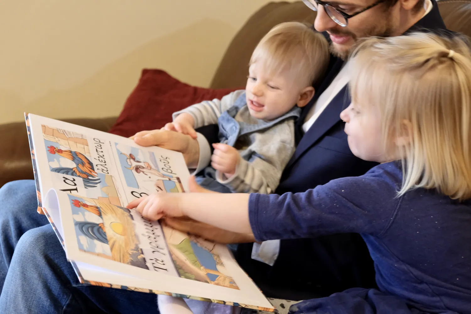 A father reading the illustrated Greek alphabet book with his two children, one pointing at the letters
